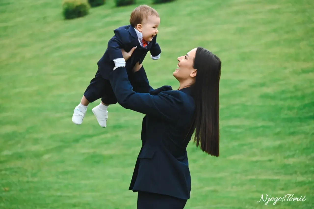 Mama u tamnoplavom odijelu podiže nasmijanu bebu u odijelu iznad glave na zelenoj livadi