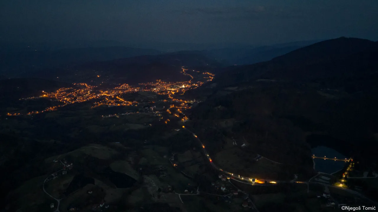 Noćni snimak dronom: osvijetljen grad u dolini, put s uličnom rasvjetom i jezero s mostom među tamnim brdima