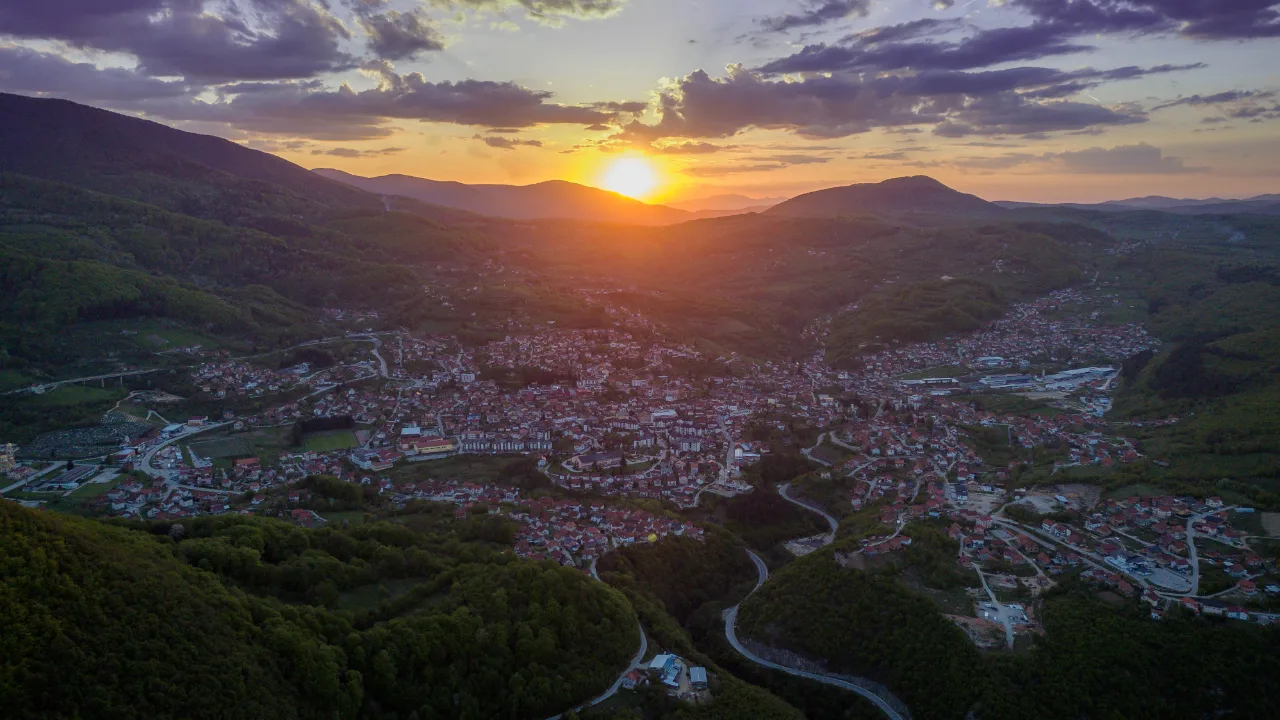 Zalazak sunca iznad planina, snimak dronom gradića u šumovitoj dolini sa serpentinama i crvenim krovovima