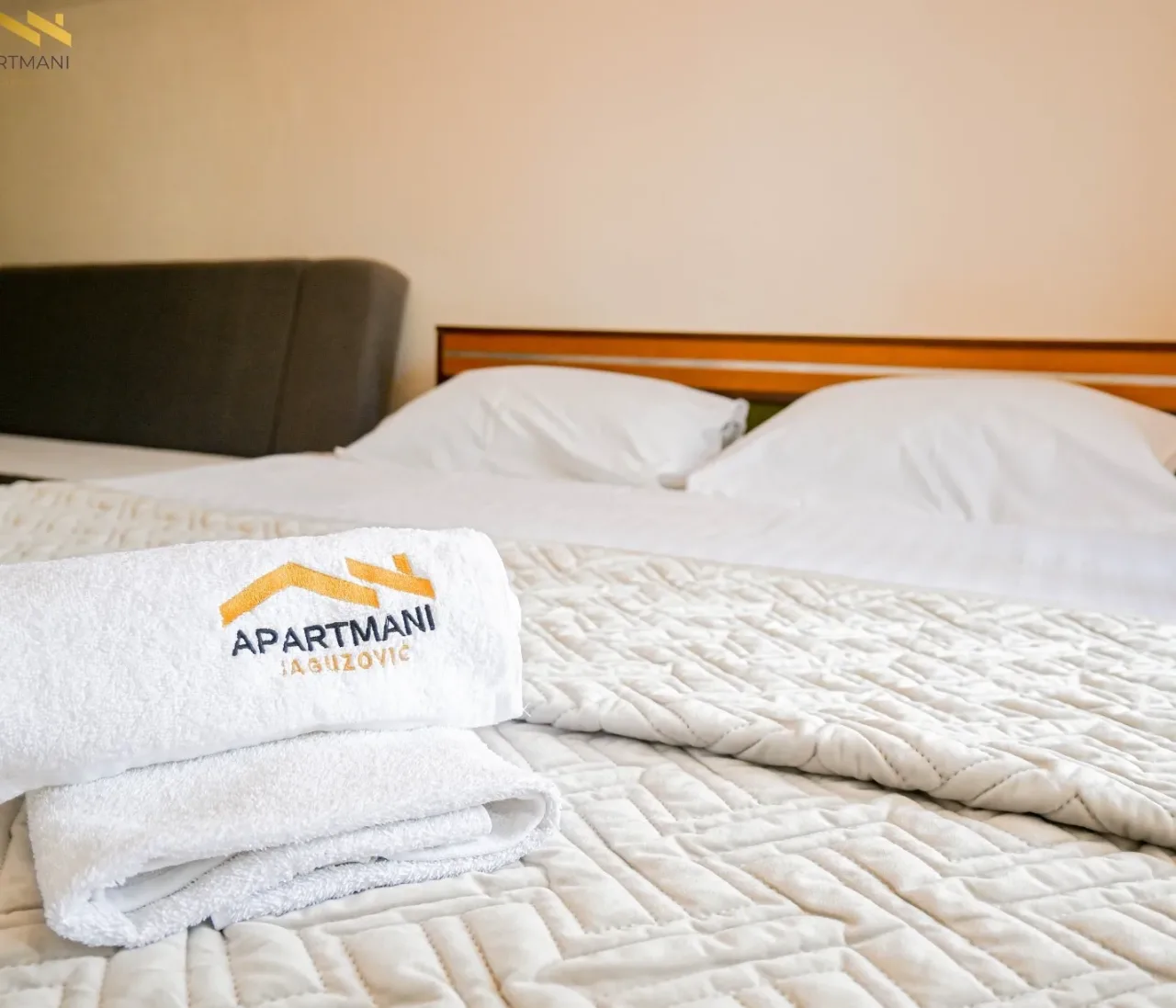 Složeni bijeli peškiri sa izvezenim logom Apartmani Jaguzović na uredno namještenom bračnom krevetu sa prošivenim prekrivačem