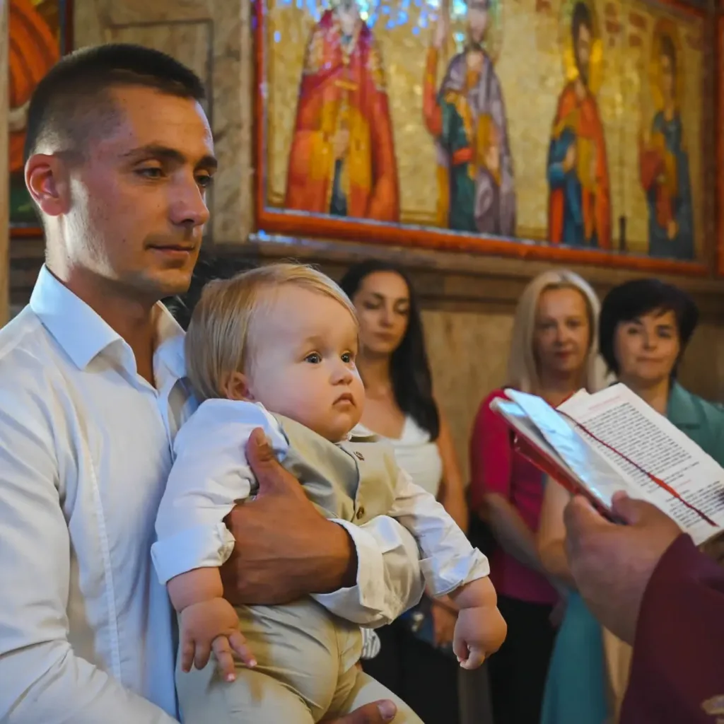 Muškarac drži dijete na krštenju u crkvi, iza njih gosti i mozaik sa likovima svetaca.