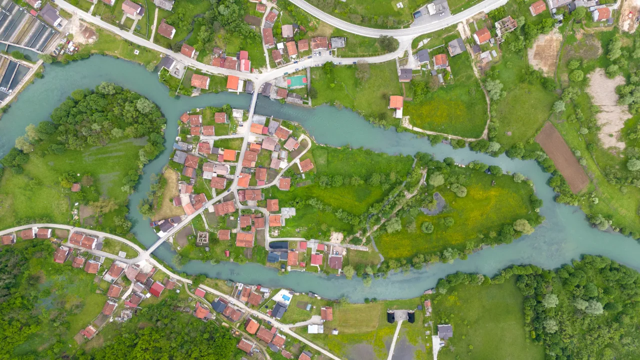 Snimak dronom odozgo: tirkizna rijeka vijuga oko naselja s crvenim krovovima, mostovi i zelena polja