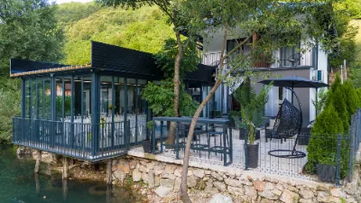 Ostakljena terasa i dvorište kuće za odmor iznad rijeke, viseća fotelja, suncobran i barski sto uz kamenu obalu