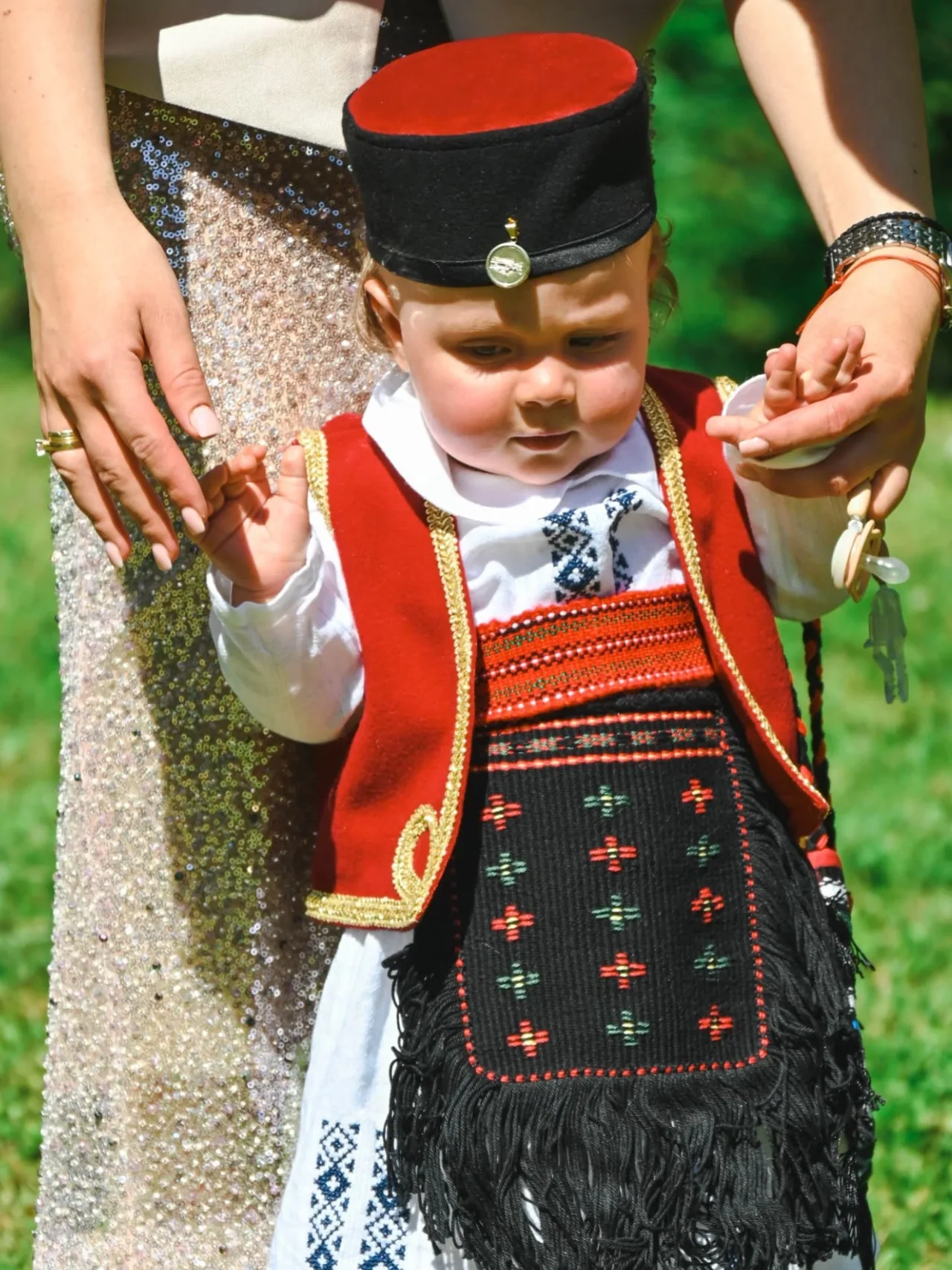 Mali dječak u narodnoj nošnji sa vezenim prslukom i kapom pravi korake dok ga odrasli drže za ruke