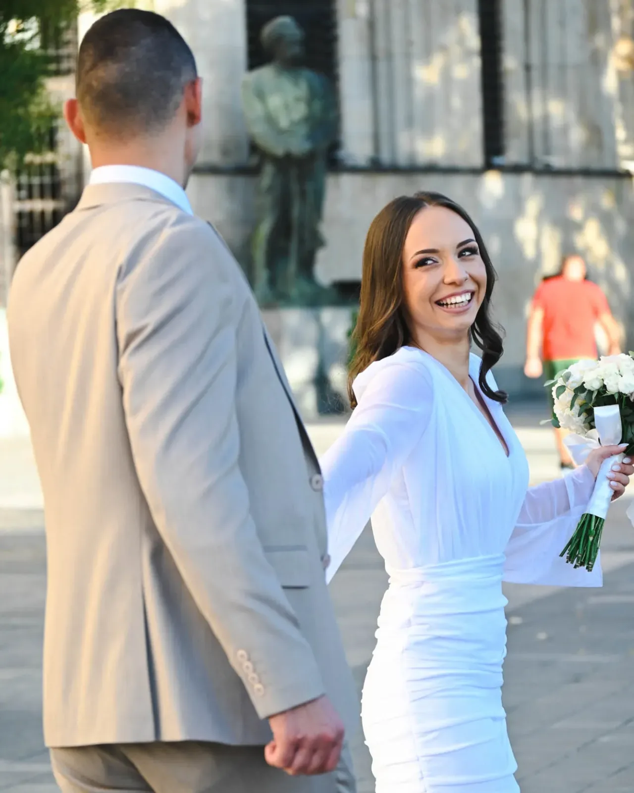 Nasmijana mlada drži mladoženju za ruku i osvrće se dok šetaju gradskim trgom, u ruci buket bijelog cvijeća