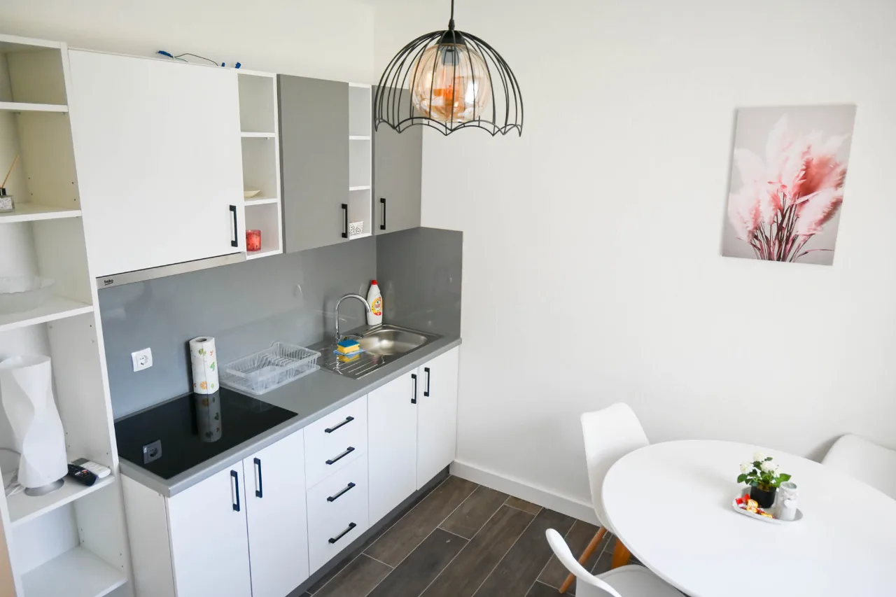 Moderna bijela kuhinja u apartmanu sa sivim elementima, staklokeramičkom pločom i okruglim stolom sa bijelim stolicama