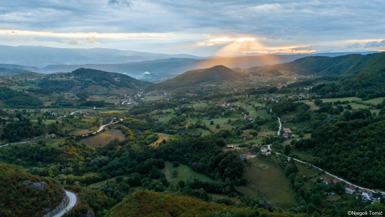 Snimak iz vazduha brdovitog kraja u jesen: sela, vijugavi putevi i zraci sunca kroz oblake iznad planina