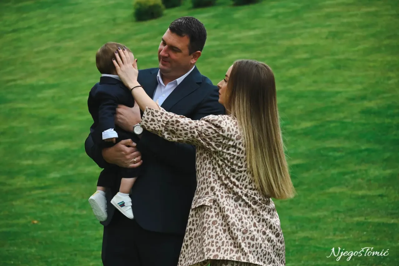 Otac u tamnom odijelu drži sina u naručju dok ga mama nježno miluje po glavi, porodični trenutak na travnjaku