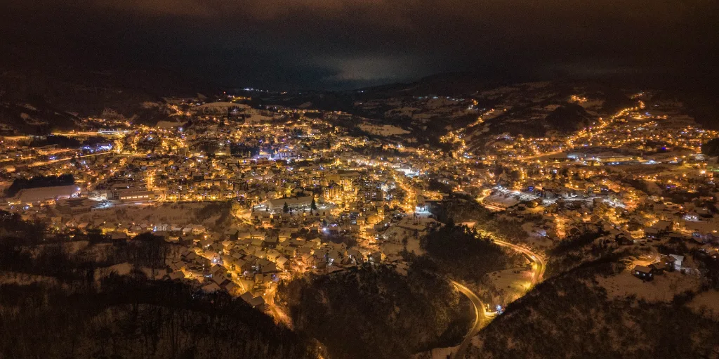 Grad noću pod snijegom, snimljen iz vazduha: osvijetljene ulice i kuće ispunjavaju dolinu, a put vijuga oko šumovitog brda.