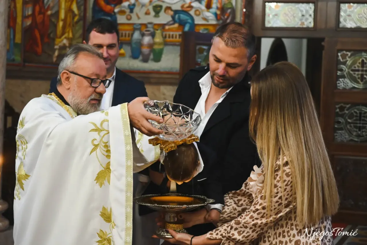 Sveštenik u bijeloj odeždi drži kristalnu posudu nad pozlaćenim pladnjem koji drži žena, obred u pravoslavnoj crkvi