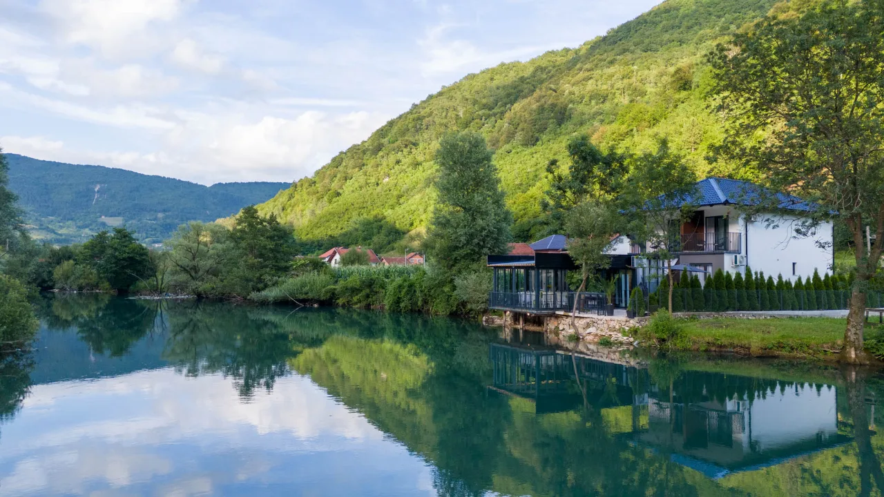 Mirna rijeka ogleda zelena brda, oblake i kuću za odmor sa ostakljenom terasom na obali, ljetni dan