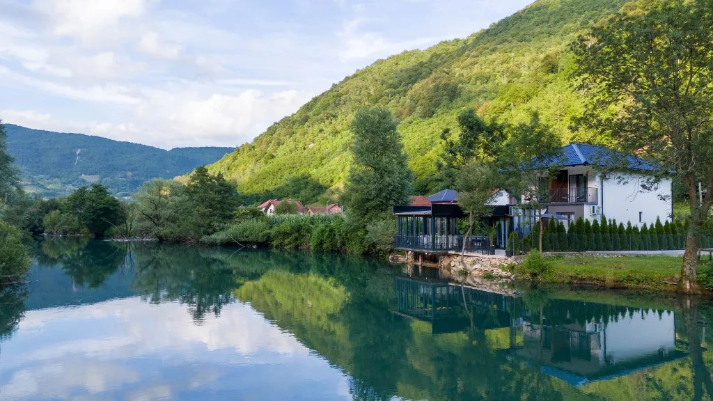 Kuća sa terasom uz rijeku, brda i nebo se ogledaju u mirnoj vodi.