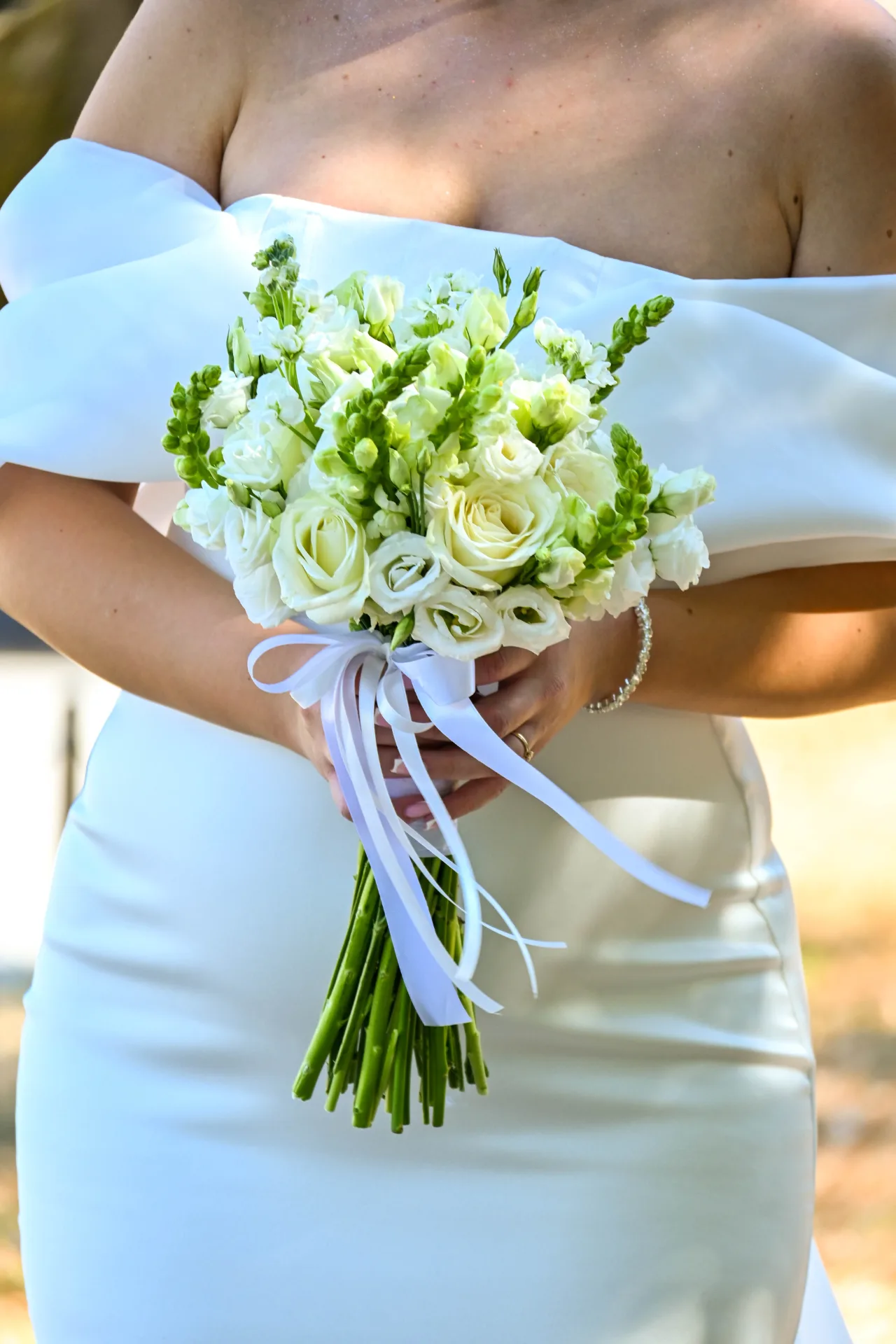 Detalj s vjenčanja: mlada u haljini spuštenih ramena drži buket bijelih ruža i zelenog cvijeća vezan trakama