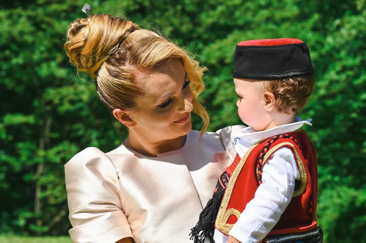 Žena u svijetloj svečanoj haljini s osmijehom gleda dječaka u narodnoj nošnji sa crvenim prslukom i kapom