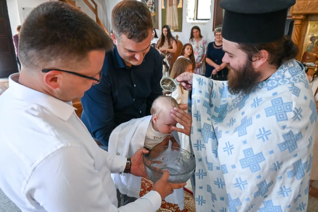 Sveštenik krštava dijete, porodica okupljena oko krstionice.
