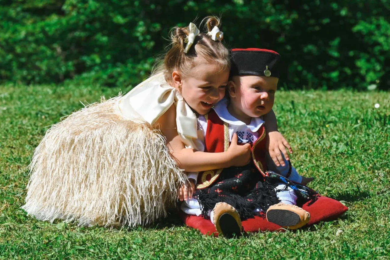 Nasmijana djevojčica sa mašnama u kosi grli bebu u narodnoj nošnji dok sjede na travi po sunčanom danu