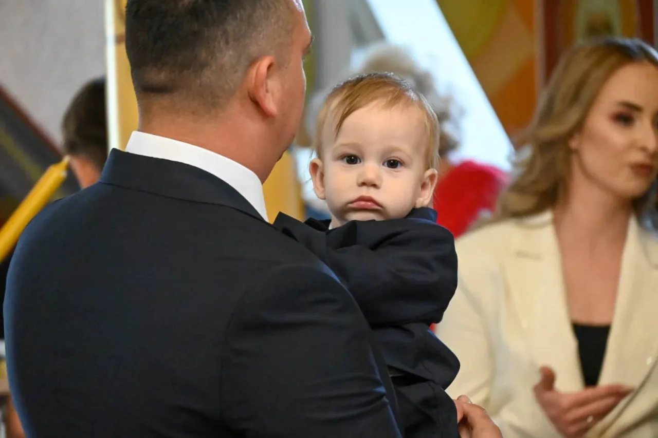 Dječak u tamnom odijelu gleda preko ramena muškarca koji ga drži u crkvi, žena u svijetlom sakou u pozadini