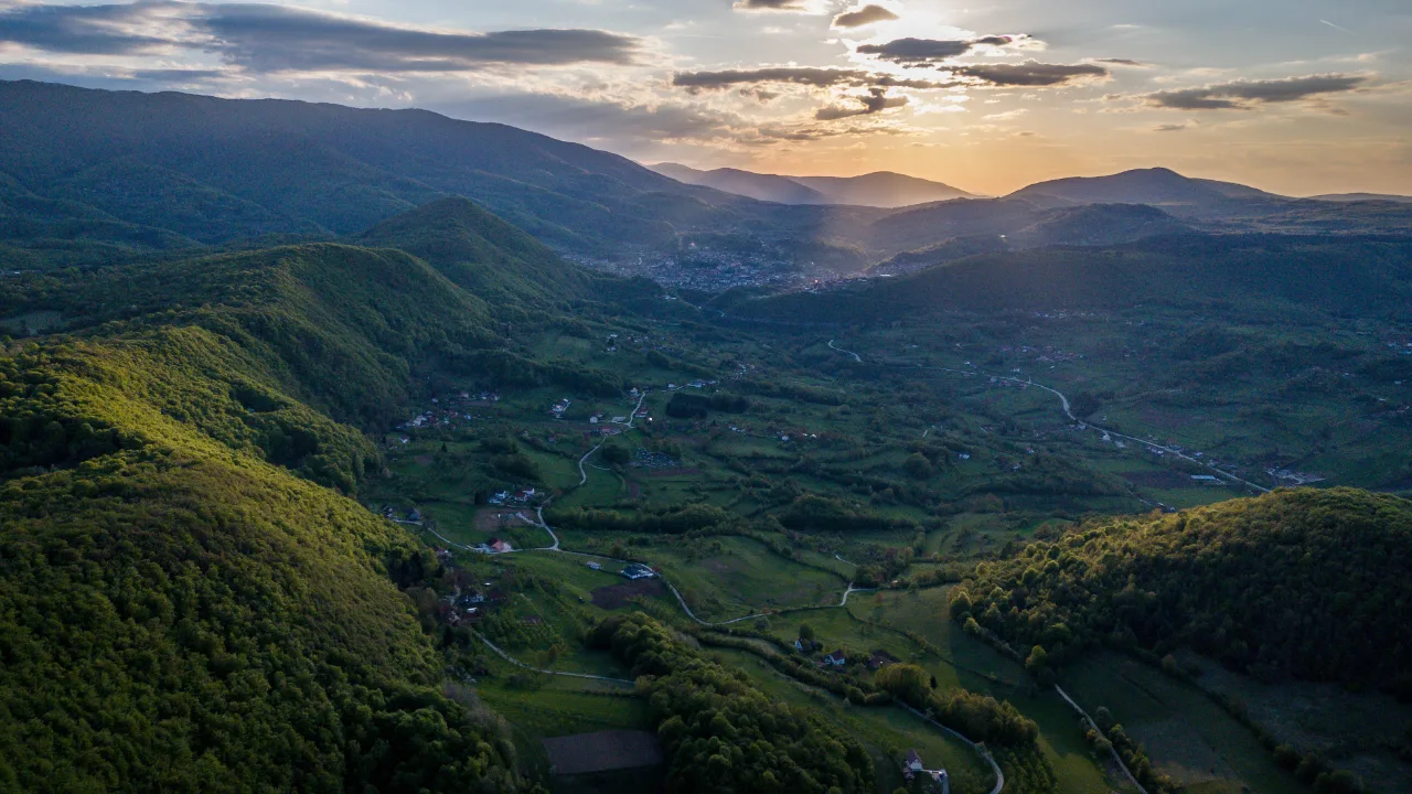 Snimak iz vazduha zelene doline u proljeće: livade, raštrkane kuće i putevi, grad u daljini pod večernjim nebom