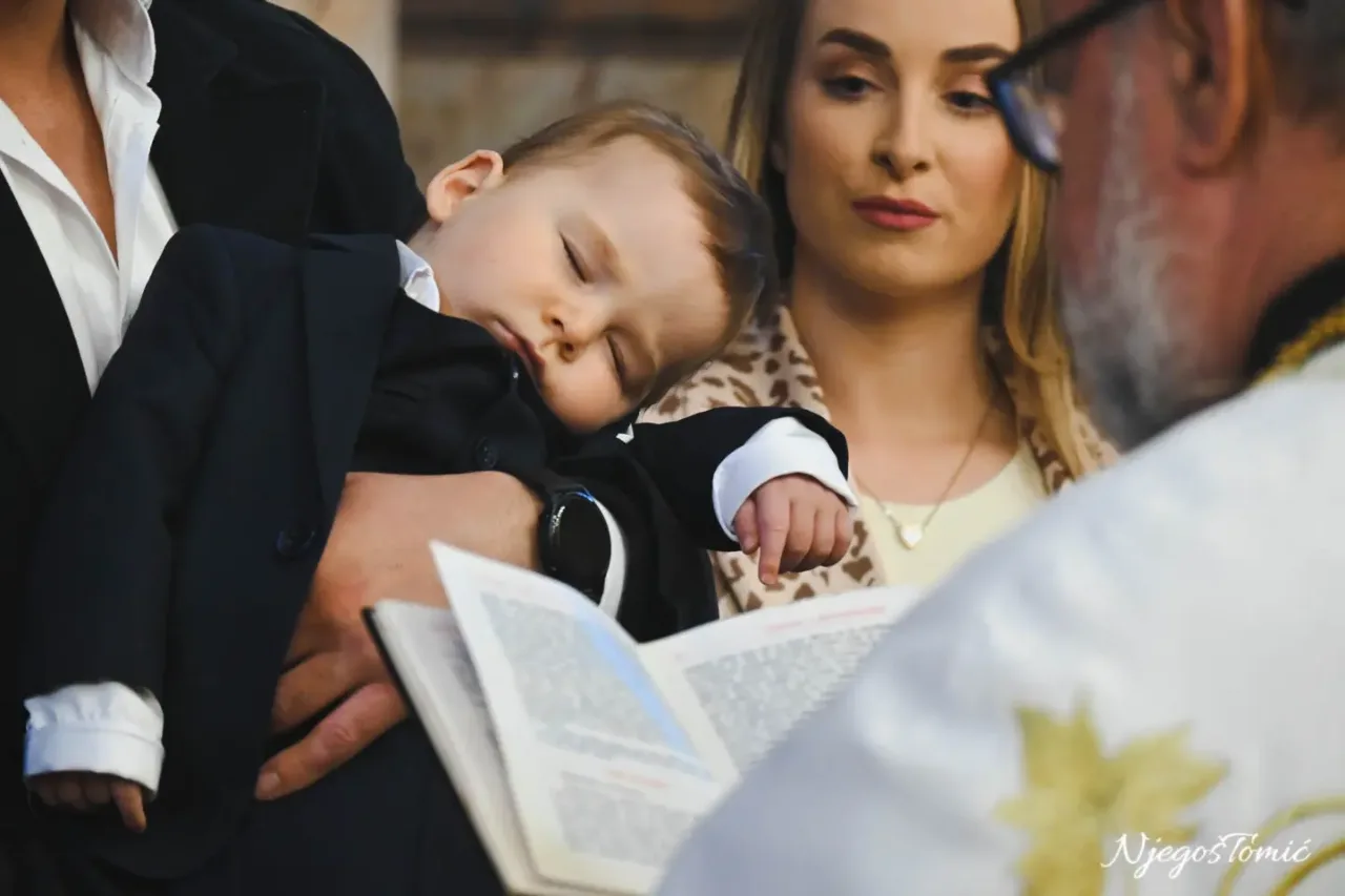 Beba u tamnom odijelu zaspala u naručju kuma dok sveštenik čita iz molitvenika, žena pažljivo prati krštenje