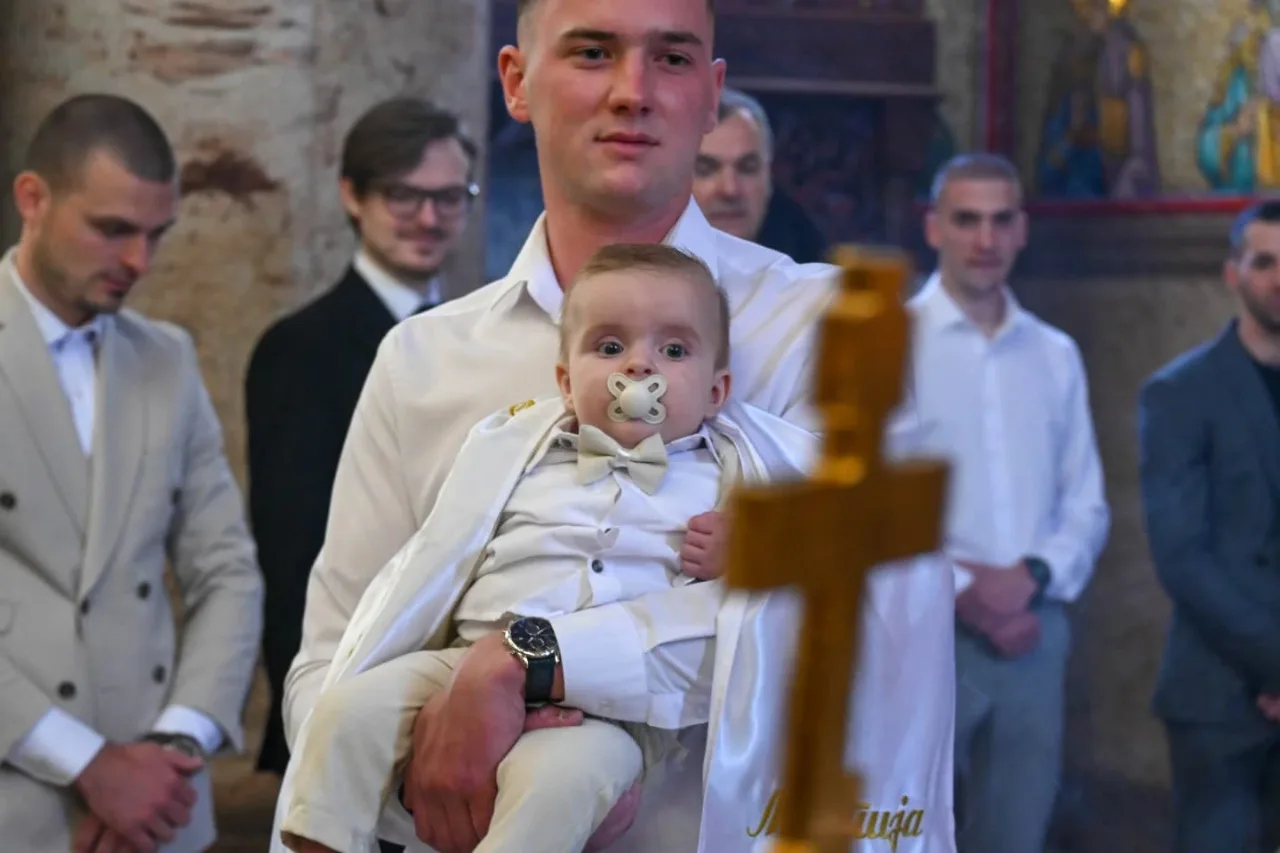 Kum drži bebu s dudom u bijelom odijelu i leptir mašni, u prvom planu zamućen pozlaćeni krst, gosti na krštenju